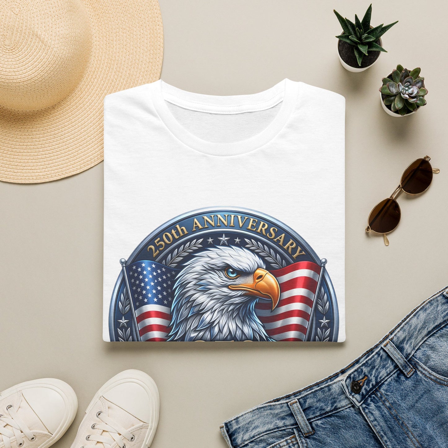 250th anniversary eagle t-shirt in white, folded flat lay with a sun hat, sunglasses and plants
