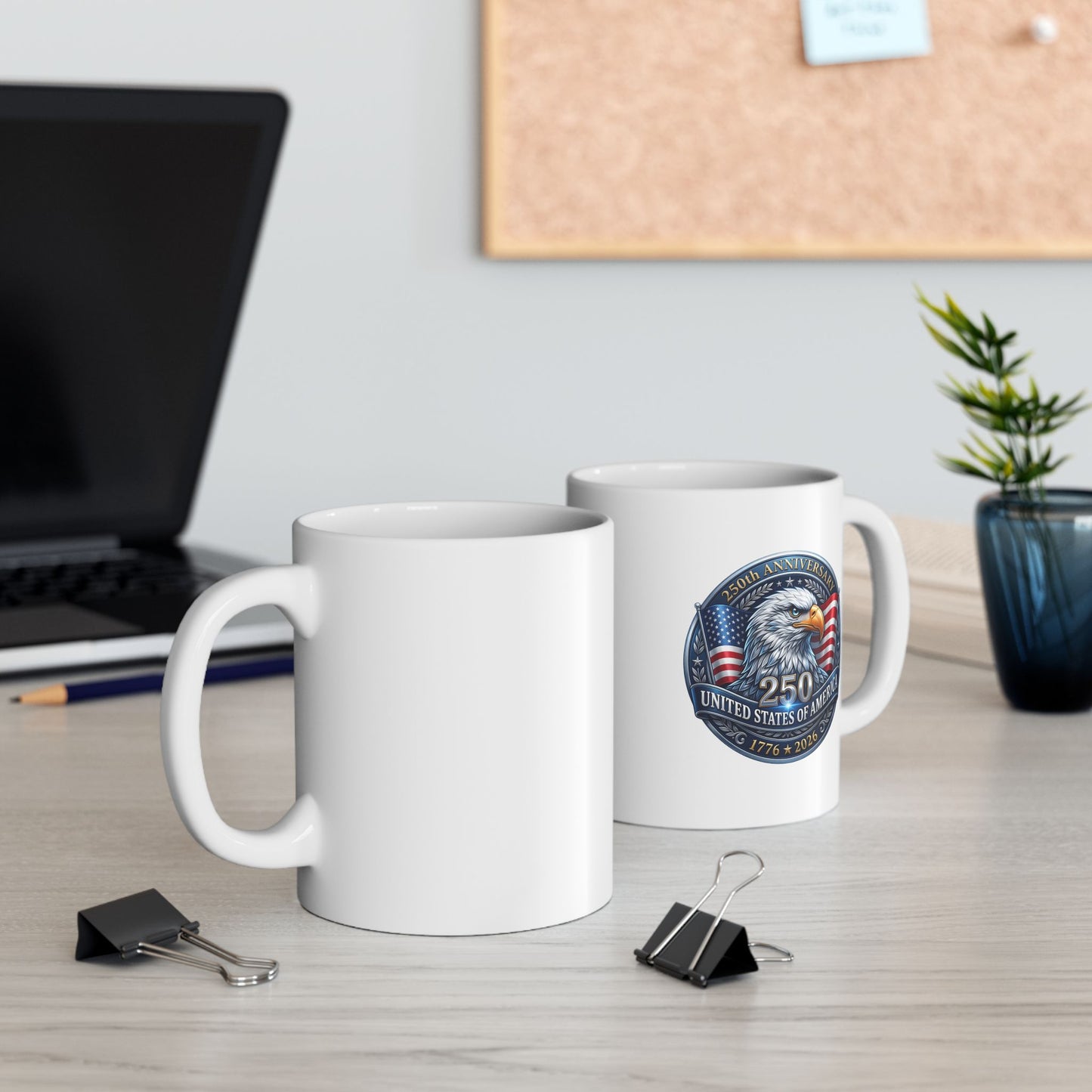 Two white eagle 250th anniversary mugs on an office desk with a laptop and corkboard, showing front and back