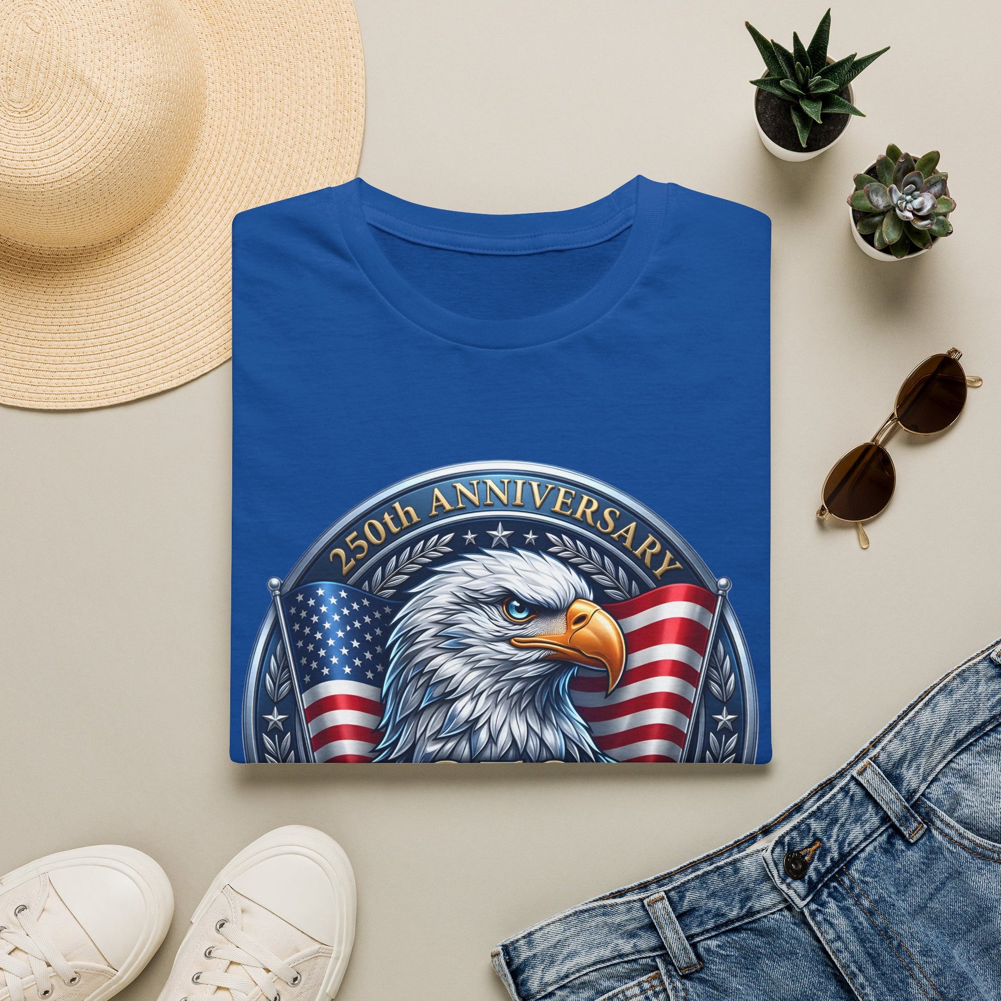 250th anniversary eagle t-shirt in blue, folded flat lay with a sun hat, sunglasses and plants