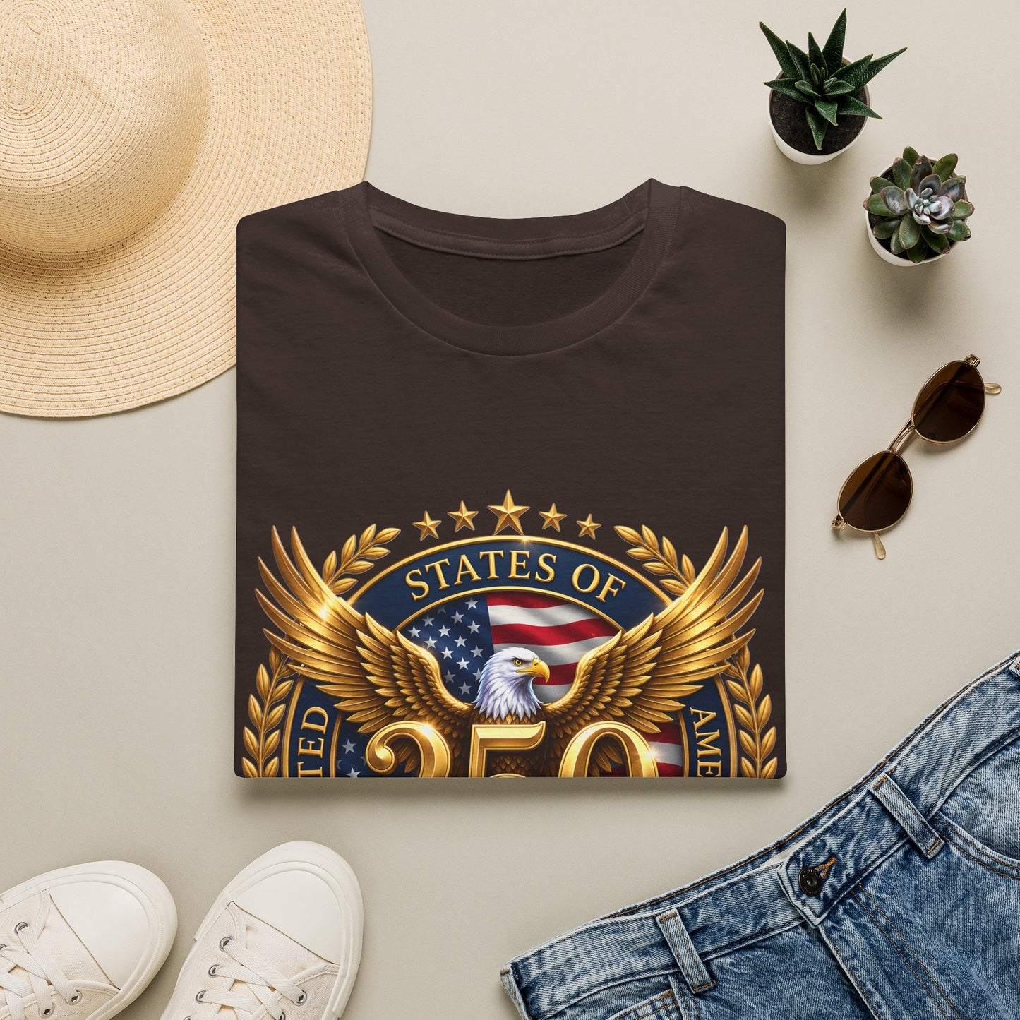 250th Anniversary USA gold eagle crest design on a dark brown t-shirt, folded flat lay with a sun hat, plants and jeans