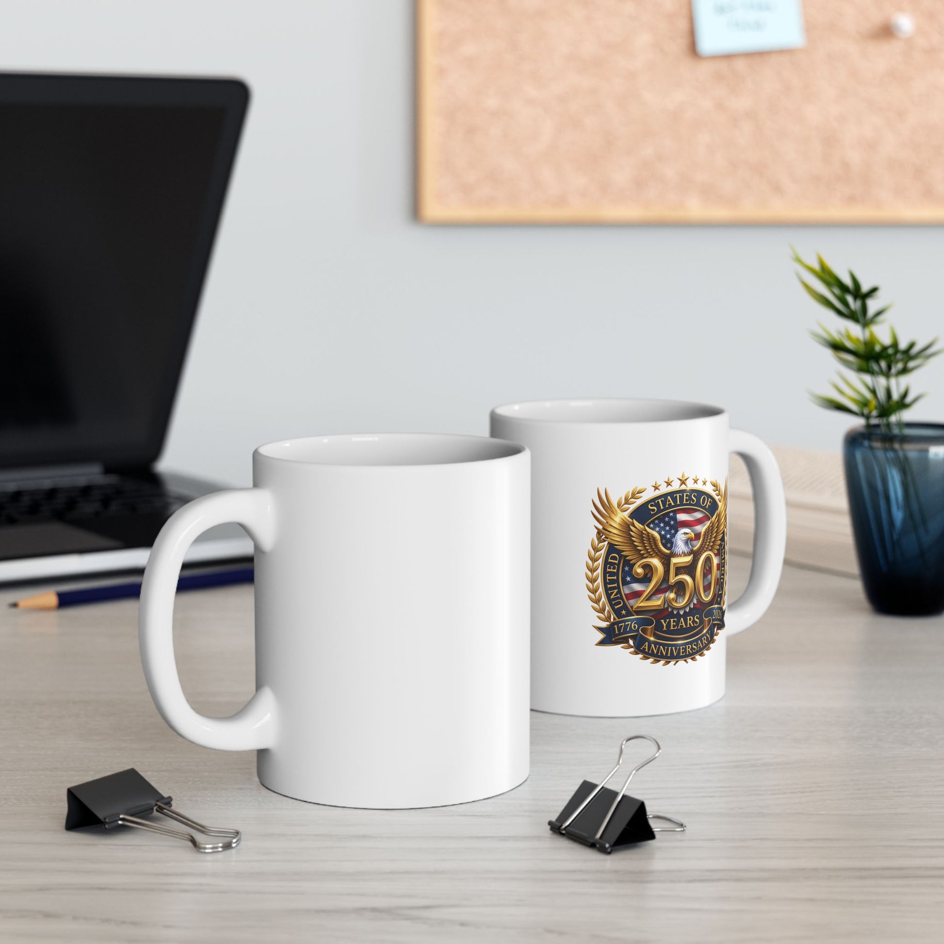 Two white ceramic mugs with the 250th anniversary eagle crest on an office desk beside a laptop and cork board